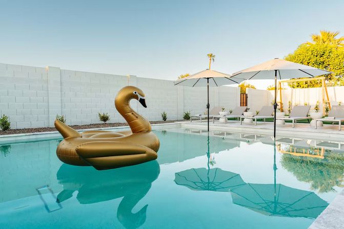 Golden swan pool float drifting in a crystal-blue backyard pool, with white lounge chairs, umbrellas and palm trees beside a modern white block wall under a clear sky.