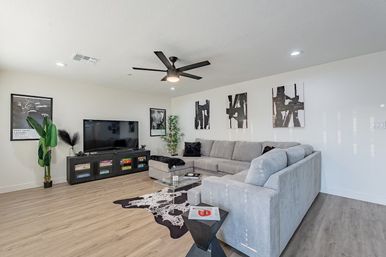 Bright modern living room with large gray sectional, glass coffee table on a black-and-white cowhide rug, flat-screen TV on a low media console, black ceiling fan, abstract black-and-white wall art, potted plants and light wood floors.