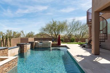 Sunny Southwestern desert backyard with a clear tiled swimming pool, stacked-stone water features, red umbrella, spiral staircase, and desert trees under a blue sky