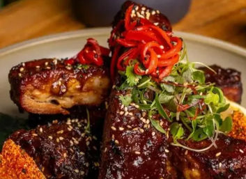 Close-up of sticky glazed pork ribs stacked on a plate, sprinkled with sesame seeds and topped with pickled red pepper curls and fresh microgreens — glossy, savory barbecue presentation.