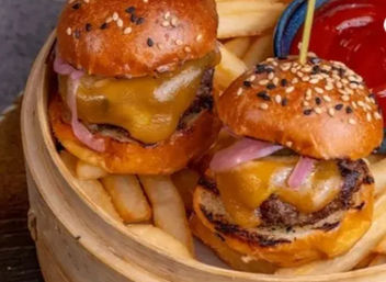 Two cheddar-topped cheeseburger sliders on sesame brioche buns with pickled red onions, served with fries and ketchup in a bamboo basket