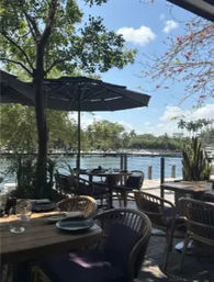 Inviting waterfront patio dining with wooden tables, wicker chairs and umbrella overlooking a calm marina and tree-lined shore on a sunny day