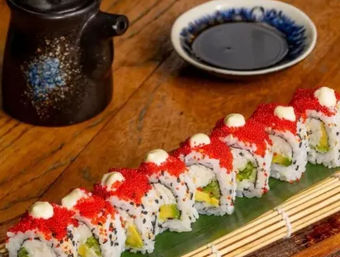 Row of colorful sushi rolls topped with red tobiko and mayonnaise dots, filled with avocado and cucumber, served on a green leaf and bamboo mat with a soy sauce dish and ceramic pourer on a wooden table.