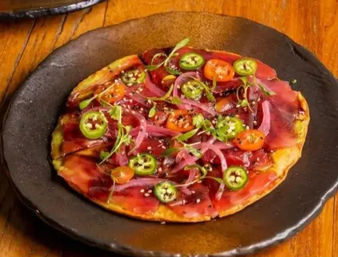 Gourmet tuna carpaccio flatbread topped with thin tuna slices, sliced jalapeños, cherry tomatoes, pickled red onions, microgreens and sesame seeds on a black ceramic plate
