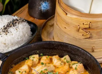 Asian-style seafood in a cast-iron skillet with creamy orange sauce, served with steamed bao in a bamboo steamer and sesame-topped white rice.