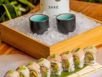 Fresh sushi roll pieces with avocado and wasabi on a bamboo mat, shot in front of a wooden ice-filled box holding a chilled sake bottle and two ceramic cups — Japanese sushi and sake presentation.