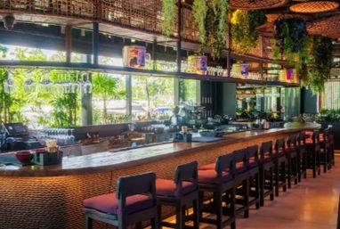 Sunlit open-air tropical-style bar interior with a long woven rattan counter, row of purple-cushioned bar stools, hanging plants and pendant lights, and large windows showing palm greenery outside