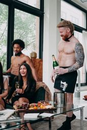 Sunlit downtown loft bachelorette party with shirtless bow-tie servers pouring champagne for laughing guests gathered around a charcuterie board.