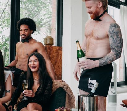 Sunlit downtown loft bachelorette party with shirtless bow-tie servers pouring champagne for laughing guests gathered around a charcuterie board.
