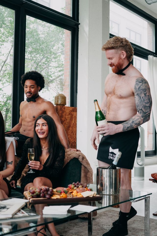 Sunlit downtown loft bachelorette party with shirtless bow-tie servers pouring champagne for laughing guests gathered around a charcuterie board.