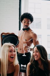 Shirtless male server in a black bow tie and apron hands a champagne flute to two laughing women in a sunlit urban loft, playful celebration vibe