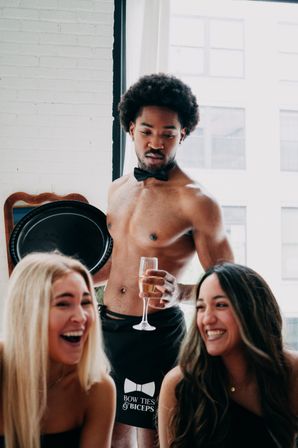 Shirtless male server in a black bow tie and apron hands a champagne flute to two laughing women in a sunlit urban loft, playful celebration vibe