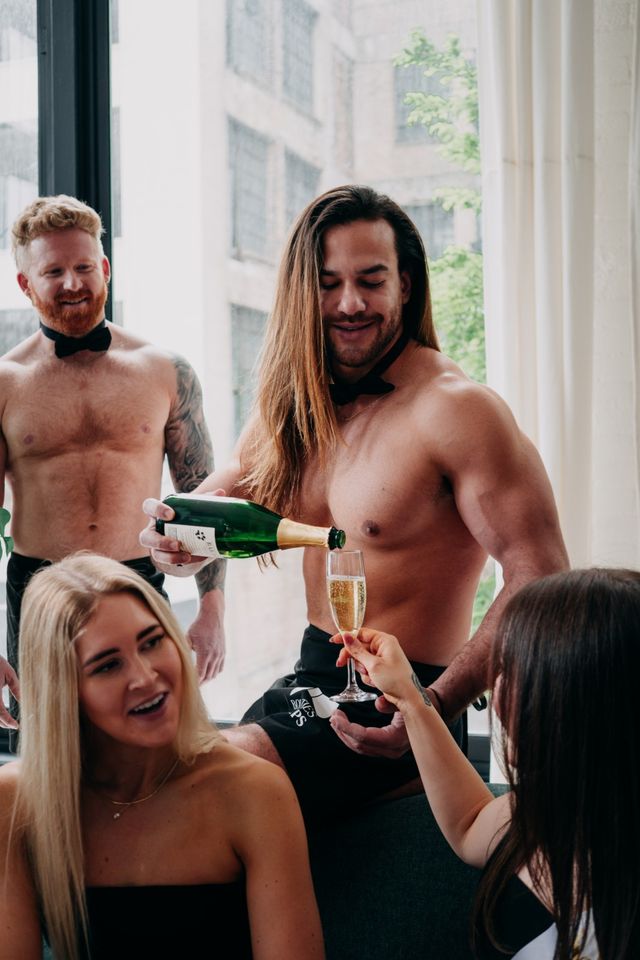 Shirtless male servers wearing bow ties pour champagne for smiling women at a bright city loft party