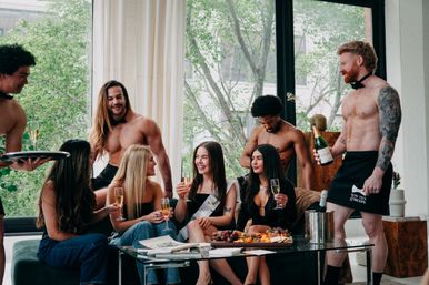 Bachelorette party in a bright loft: sash-wearing guest and friends clinking champagne glasses while shirtless servers pour drinks and a charcuterie board sits on the coffee table.
