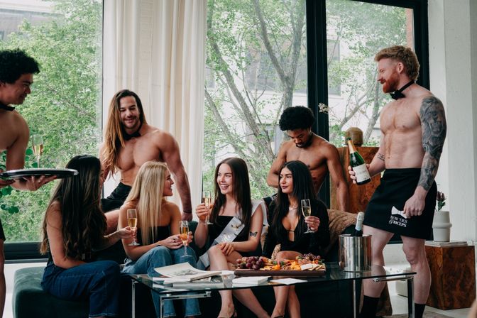 Bachelorette party in a bright loft: sash-wearing guest and friends clinking champagne glasses while shirtless servers pour drinks and a charcuterie board sits on the coffee table.