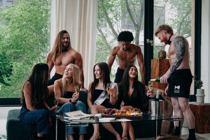 Bachelorette party in a bright urban loft: bride-to-be wearing a sash and friends laughing on a sofa while shirtless servers pour champagne beside a charcuterie board on a glass coffee table.