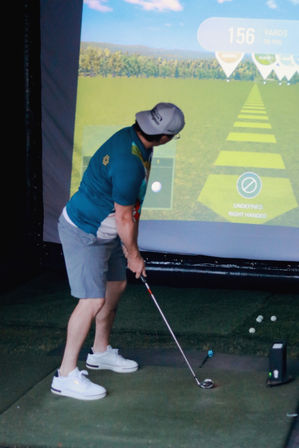 Golfer practicing a swing on an indoor golf simulator, standing on a turf mat in shorts and white sneakers, aiming toward an on-screen 156-yard target with golf club and scattered balls nearby.