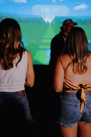 Two people in summer tops and denim shorts stand with their backs to camera in an indoor golf simulator, watching a projected virtual fairway and distance marker — casual indoor golf entertainment scene.