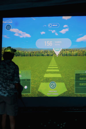 Silhouette of a golfer at an indoor golf simulator aiming down a virtual fairway; projection shows target markers and a large “156 yards to pin” distance readout.