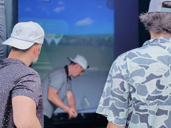 Three men wearing caps at an indoor golf simulator bay, one swinging a club toward a large screen showing a virtual fairway