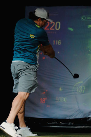 Golfer in a cap and shorts swinging a driver inside an indoor golf simulator, mid-swing with distance and angle data visible on the virtual screen.
