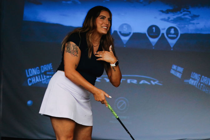 Smiling female golfer in black top and white skirt holding a driver at an indoor golf simulator with projected target graphics and 'Long Drive Challenge' text