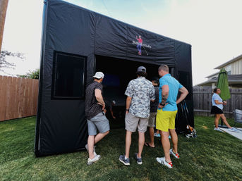 Group of people gathered around a black pop-up golf simulator tent on a suburban backyard lawn, watching gameplay inside with golf clubs and a wooden fence visible