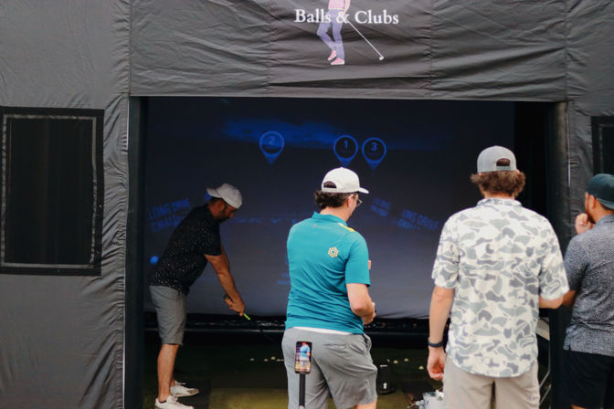 Four golfers in a covered golf-simulator bay — one player swings toward a projected target screen while three others watch, with a smartphone on a stand recording the session.