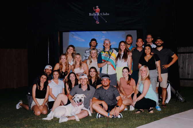 Group of smiling adults posing on a backyard lawn at an evening outdoor movie setup with a projector screen and a playful white dog in front.