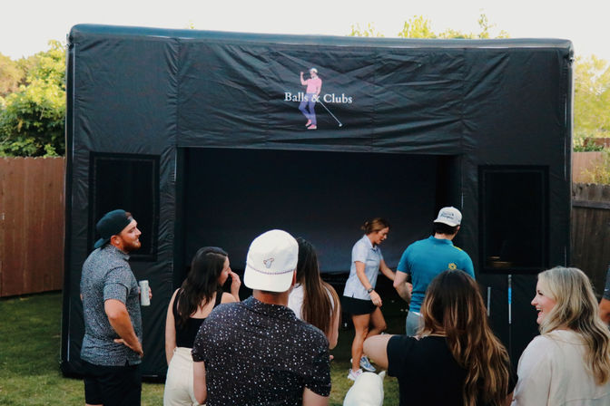 Friends gathered in a backyard around a black inflatable golf simulator tent, taking turns using a portable virtual golf setup at an outdoor party