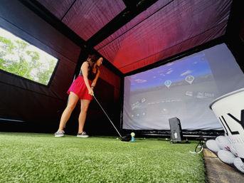 Female golfer in a red skirt teeing off inside a portable indoor golf simulator tent on artificial turf, aiming at a projected driving-range screen with a bucket of practice balls.
