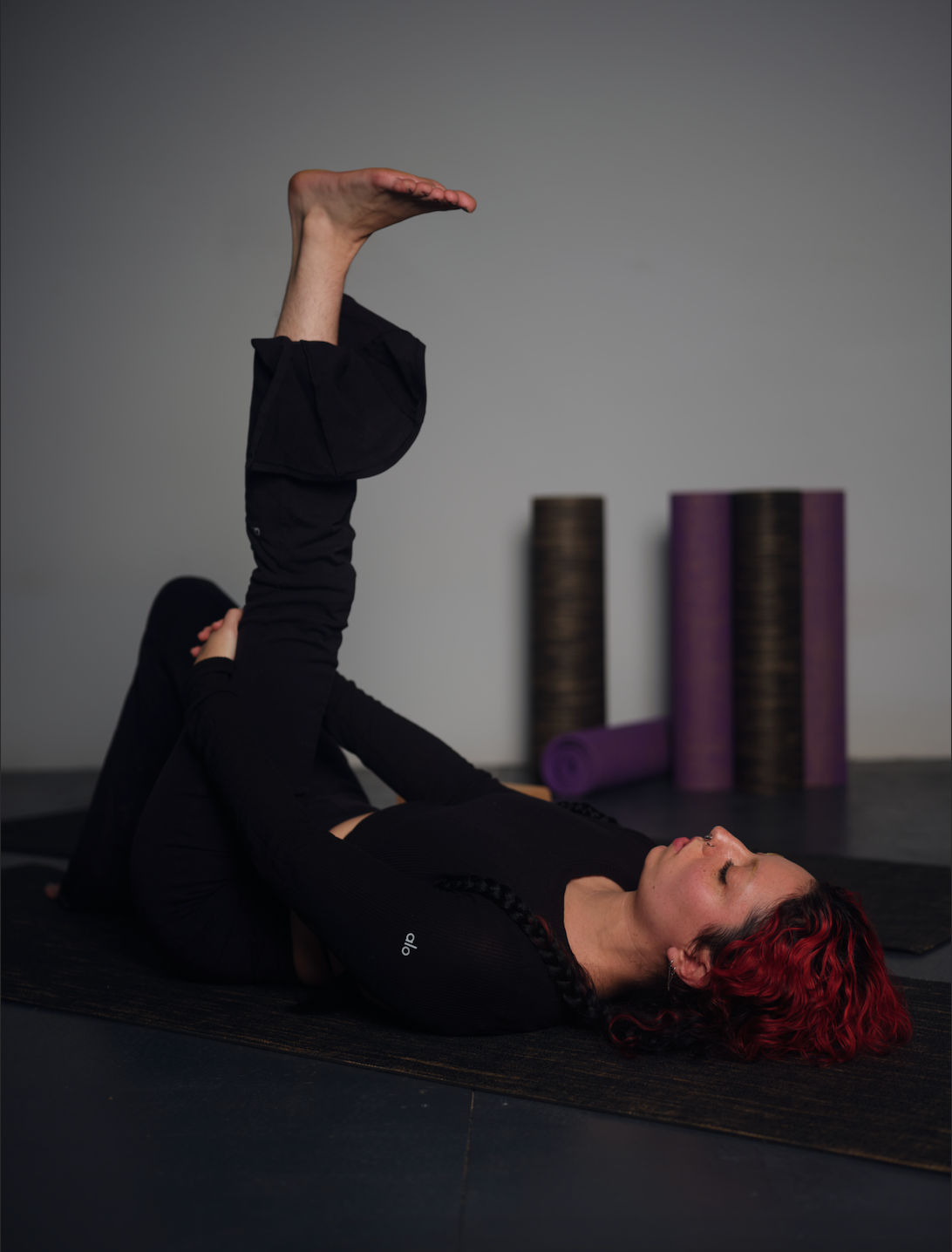 Person with red hair doing a supine hamstring stretch on a black yoga mat in an indoor yoga studio, foam rollers and purple mats visible in the background.