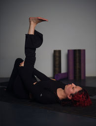 Person with red hair doing a supine hamstring stretch on a black yoga mat in an indoor yoga studio, foam rollers and purple mats visible in the background.