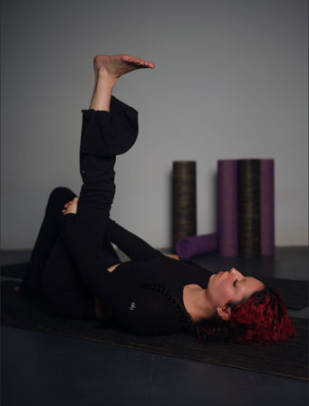 Person with red hair doing a supine hamstring stretch on a black yoga mat in an indoor yoga studio, foam rollers and purple mats visible in the background.
