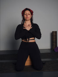 Person kneeling on a cork yoga block in a studio, hands crossed over chest and eyes closed in a calm meditation pose, wearing black activewear with a purple rolled mat and foam roller visible behind.