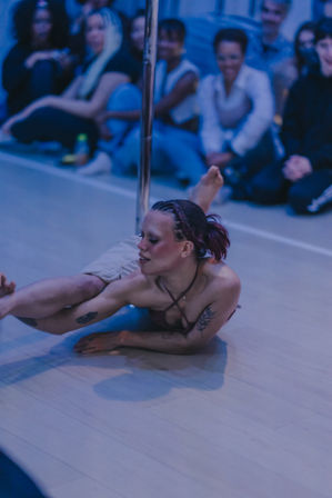 Pole dance performer in an indoor studio executing a floor move by a chrome pole under moody blue lighting, braided hair and visible tattoos, seated audience watching nearby.