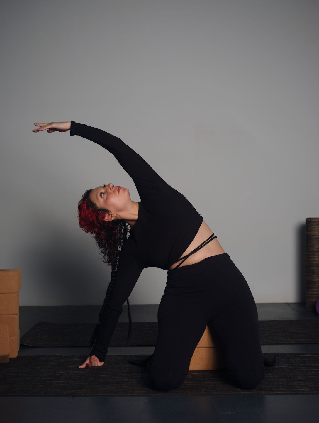 Kneeling side-stretch yoga pose: person with red hair in a black outfit reaching overhead on a mat beside cork yoga blocks in a minimalist indoor studio.