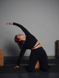 Kneeling side-stretch yoga pose: person with red hair in a black outfit reaching overhead on a mat beside cork yoga blocks in a minimalist indoor studio.