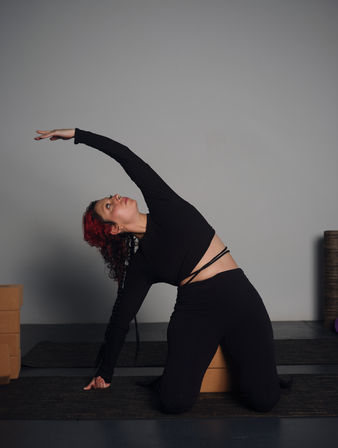Kneeling side-stretch yoga pose: person with red hair in a black outfit reaching overhead on a mat beside cork yoga blocks in a minimalist indoor studio.