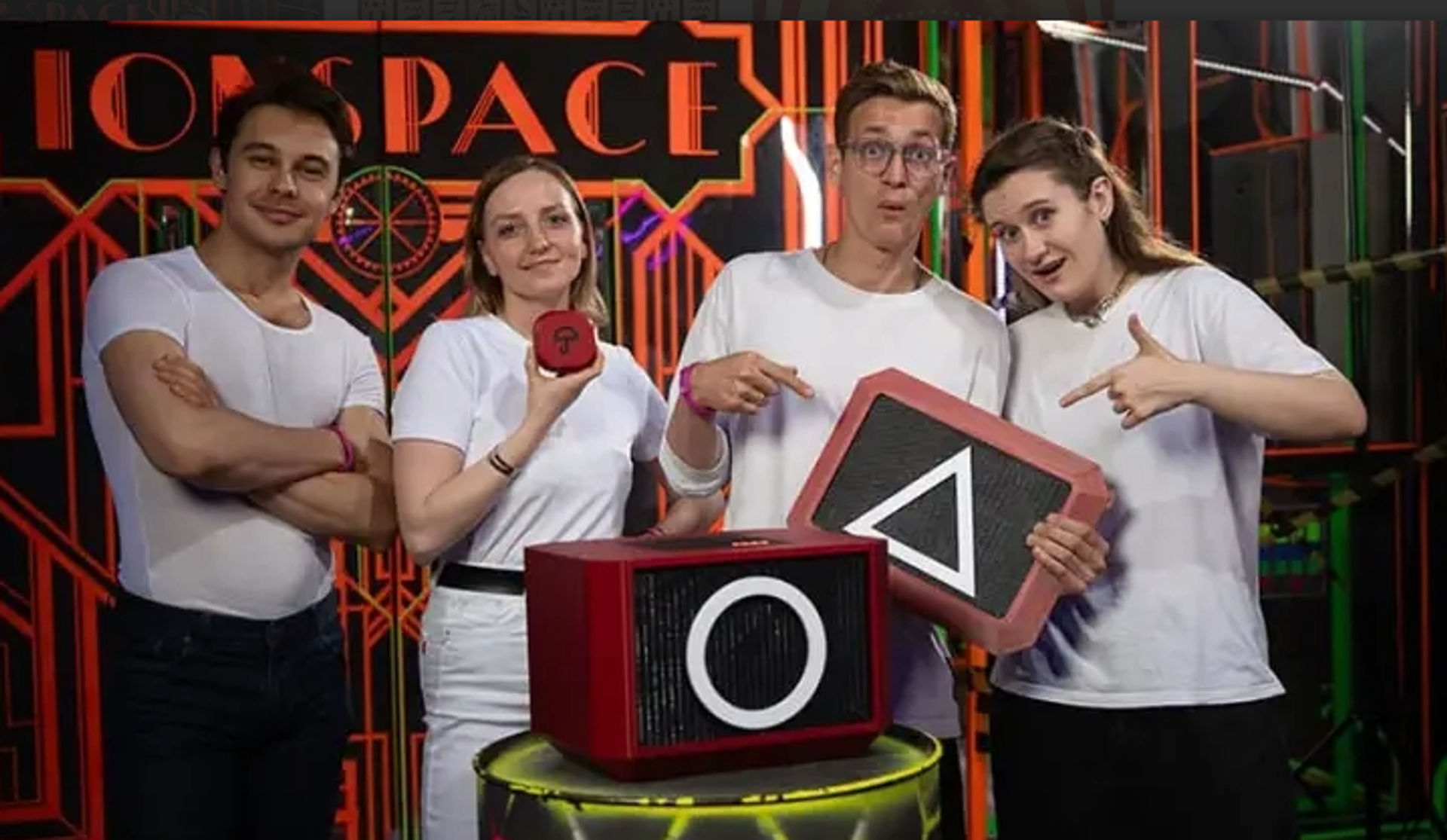 Four people in white shirts pose with red geometric game props showing a circle and triangle on a neon-lit indoor game-room set.
