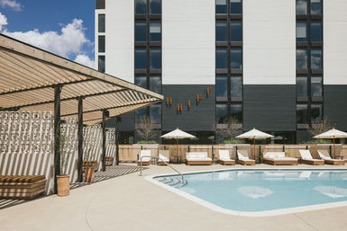Sunlit urban hotel pool deck with pergola cabanas, white umbrellas, wooden lounge beds and a modern high-rise facade.