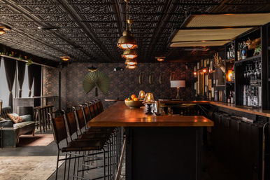Cozy, moody cocktail bar interior with a long wooden counter, row of leather high stools, industrial pendant lights, ornate tin ceiling, floral wallpaper, shelves of spirits and glassware, and a lounge seating area by curtained windows.