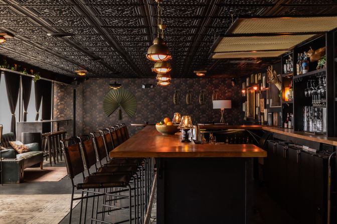 Cozy, moody cocktail bar interior with a long wooden counter, row of leather high stools, industrial pendant lights, ornate tin ceiling, floral wallpaper, shelves of spirits and glassware, and a lounge seating area by curtained windows.