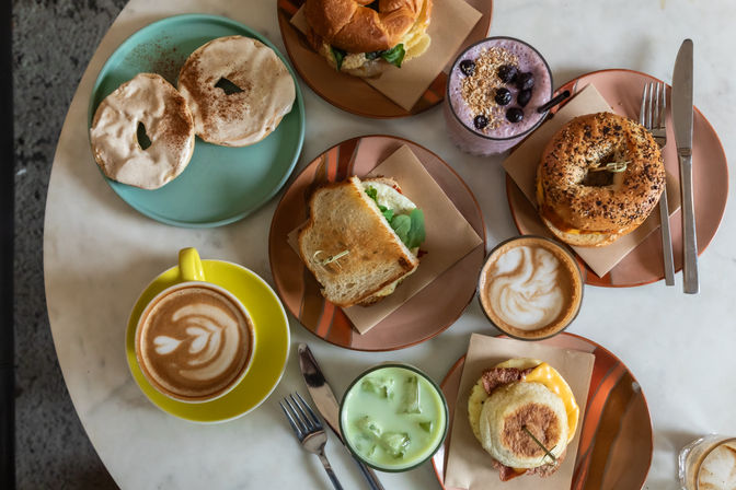 Top-down brunch spread on a marble cafe table — bagel sandwiches, toasted sandwich, egg-and-cheese English muffin, blueberry smoothie, iced matcha, and two lattes with latte art on colorful plates.