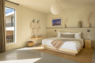 Sunlit minimalist bedroom with a low wood platform bed, neutral linens and throw, floating shelf holding two hanging robes, woven rug, pendant light and a large window revealing an exterior balcony.