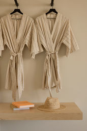 Two beige striped cotton robes on black hangers against a sandy wall above a light wood shelf holding a woven straw sun hat and two stacked books — minimalist coastal spa dressing nook.