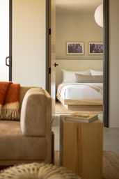 Peek-through view from living room into a cozy modern apartment bedroom with white linens, beige throw, wooden platform bed, framed art, and neutral minimalist decor.