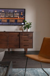 Mid-century modern living room with wall-mounted TV, wooden media console topped with a potted plant and book, tan leather lounge chair on a blue area rug and dark hardwood floors.