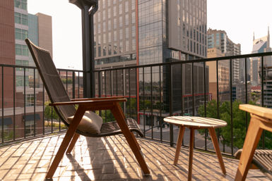 Sunlit urban balcony with wooden lounge chairs and a small round table overlooking downtown high-rise buildings and a tree-lined city street.