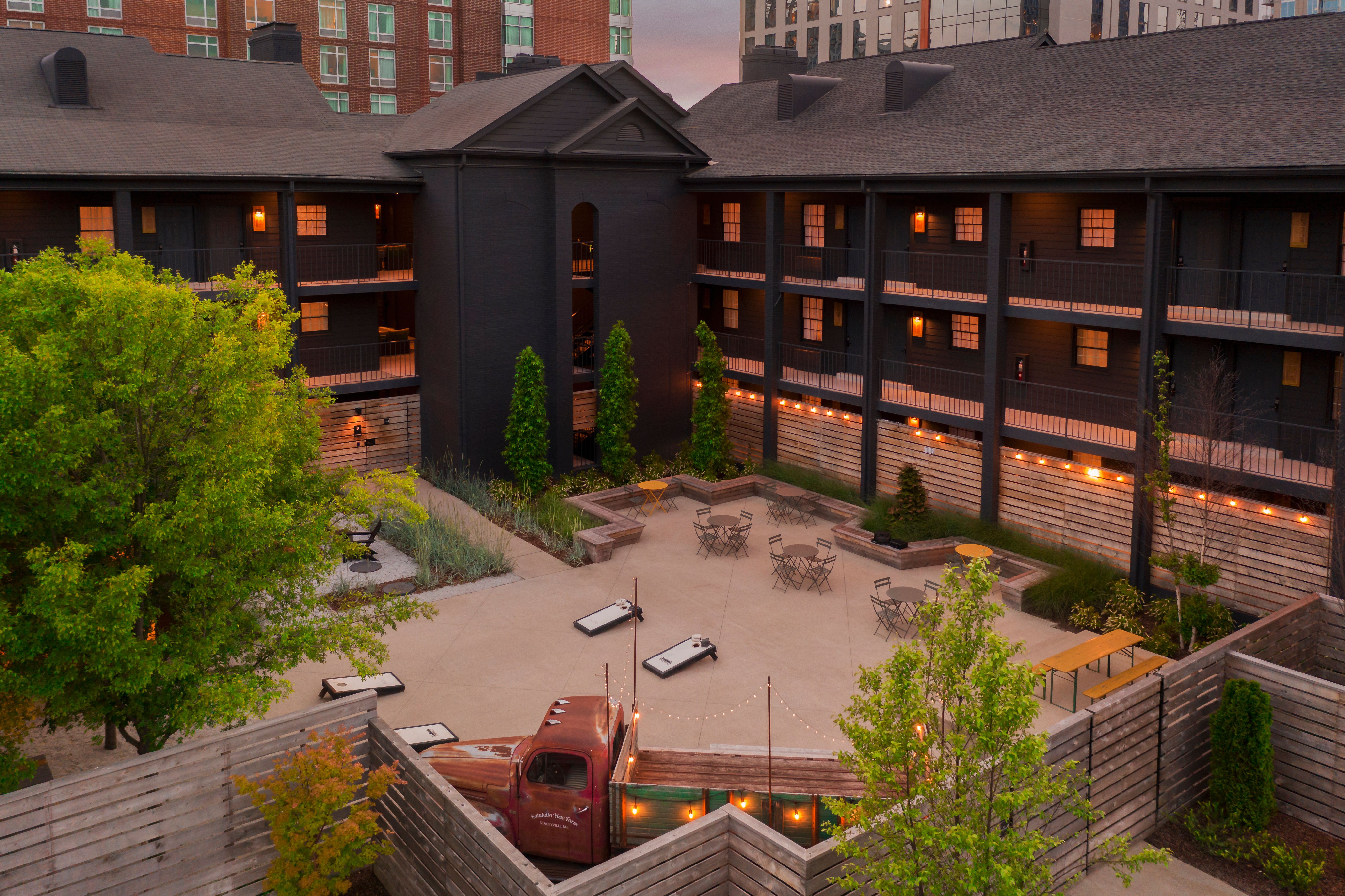Urban hotel courtyard at dusk with string lights, patio tables, cornhole boards and a vintage truck centerpiece, surrounded by dark wood three-story balconies and city buildings.